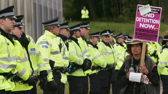Mujer con pancarta de protesta que reza "fin a la detención ahora" frente a una fila de policías a la entrada del centro de internamiento de extranjeros de Dungavel, en Escocia