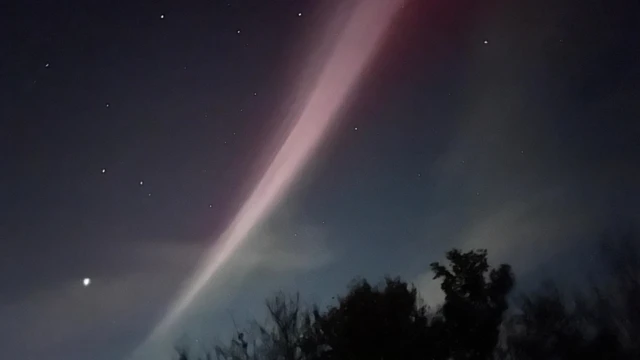 Thin stream of purple and white light in the night sky with some white cloud and bright stars.