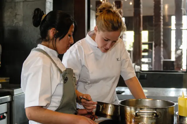 Chef le muestra el contenido de una olla a una estudiante de cocina