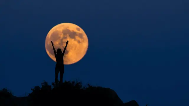 Uma lua cheia laranja, com uma mulher na frente dela, os braços no ar