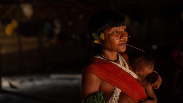 Mulher yanomami, com adereçosblaze apostas como jogarpenas e madeira no rosto, carrega bebêblaze apostas como jogarseu colo