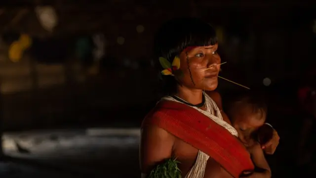 Mulher yanomami, com adereçosgestão de banca betfairpenas e madeira no rosto, carrega bebêgestão de banca betfairseu colo