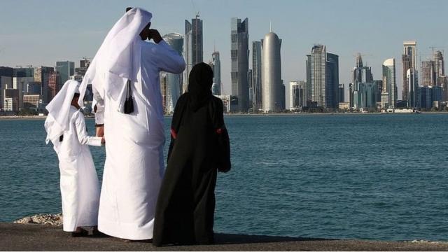 A man and two children in traditional dress stare across water at a city in the Persian Gulf