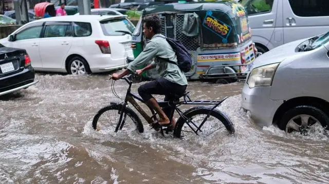 ঢাকায় ভারী বর্ষণের পর, জলমগ্ন রাস্তার মধ্য দিয়ে যাচ্ছে বিভিন্ন যানবাহন। একটি সাইকেলে করে যাচ্ছেন একজন ব্যক্তি, সাইকেলটির চাকাগুলো প্রায় অর্ধেক পানিতে ডুবে আছে