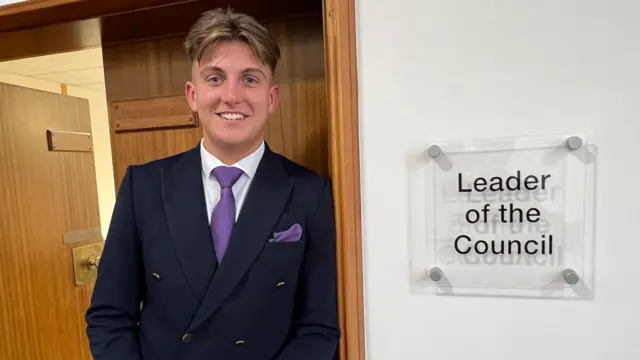 George Finch, con traje azul y corbata morada, se apoya en la puerta de la oficina del líder del consejo. El cartel a su izquierda dice "Líder del Consejo".