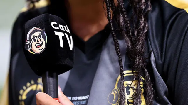 A imagem mostra um enquadramento fechado de um microfone com a marca CazéTV sendo segurado por uma mulher vestindo uma camisa preta com o logotipo do canal. O rosto da pessoa não aparece, apenas parte do pescoço, do cabelo trançado e do torso.