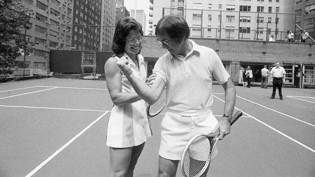 Czarno-białe zdjęcie przedstawiające Billie Jean King dotykającą bicepsa Bobby'ego Riggsa. Są na korcie tenisowym i obaj trzymają rakiety.