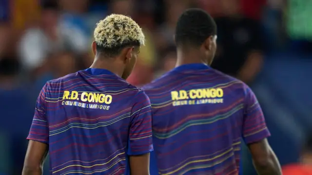 On voit de dos les joueurs du FC Barcelone Lamine Yamal (à gauche) et Marcus Rashford (à droite) pendant l'échauffement avant un match. Yamal, avec ses cheveux blonds décolorés, est plus proche de l'appareil photo et parfaitement net, tandis que Rashford est légèrement flou. On voit les deux joueurs de la taille jusqu'au haut du torse, arborant leurs maillots d'entraînement violets ornés de lignes horizontales ondulées dans différentes nuances de rouge et de jaune, ainsi que la phrase « RD Congo, le cœur de l'Afrique » écrite en français en lettres jaunes dans le haut du dos.