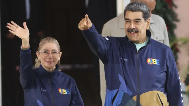 Cilia Flores saluda al parecer a una multitud con el brazo extendido. A su lado, Nicolás Maduro señala a un punto fuera del plano. Ambos sonríen.