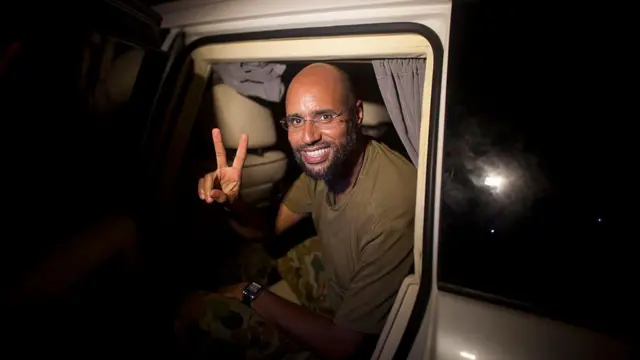 Seyfülislam Kaddafi bir aracın içinde, üzerinde asker yeşili bir t-shirt var ve sağ eliyle V şekilde zafer işareti yapıyor. Fotoğraf 15 yıl öncesine ait, 23 Ağustos 2011'de çekilmiş. 