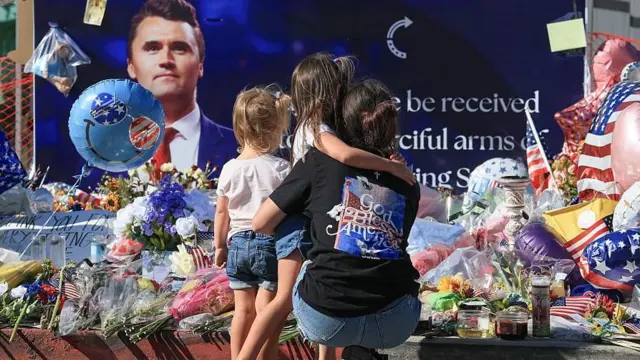 Um memorial improvisado foi montado do lado de fora da sede da Turning Point USA em Phoenix, onde uma mãe e seus dois filhos colocam flores diante de um grande pôster de Charlie Kirk.