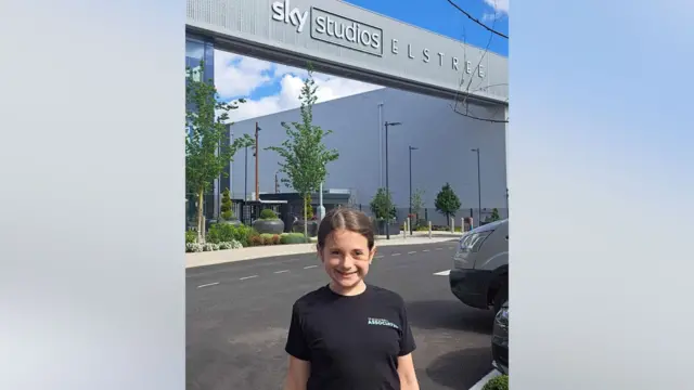 Lily está de pie frente a un gran edificio gris con el lema Sky Studios Elstree. Lleva un abrigo negro, un jersey blanco y pantalones rosas estampados.