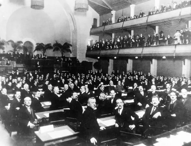 Чорно-біле фото, на якому багато чоловіків сидять у великій залі та на двох балконах з правого боку зали