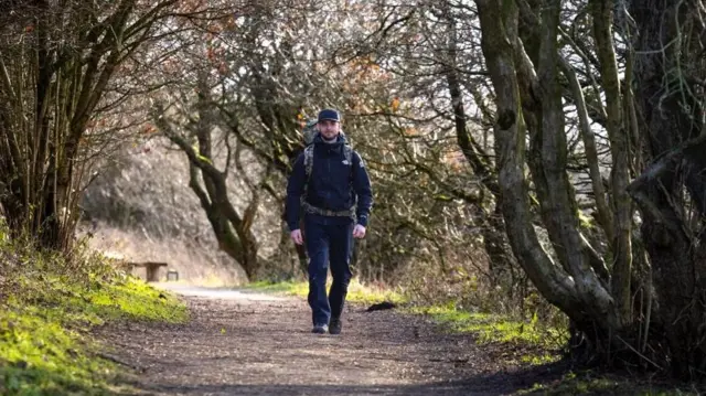 Un hombre vestido de oscuro lleva una mochila, camina por un sendero con césped y árboles a ambos lados.