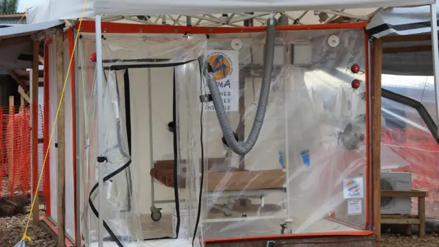 Structure en plastique transparent en forme de cube avec une porte zippée ouverte. Il y a un lit vide à l'intérieur et aucun patient