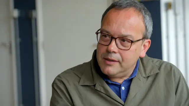 Nick Lowles, gri saçlı ve koyu çerçeveli gözlüklü, mavi gömleğin üzerine gri-yeşil bir ceket giyen bir adam