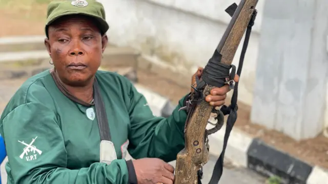 Mama Marian Alarape Iyanuore ree to wọ asọ ologun alawọ ewe, o joko, kọ ẹyin si ile kan, o si wa lori aga kan, to si gbe ibọn sakabula lọwọ.