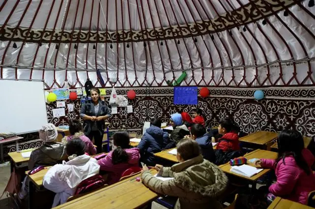 Çocukların süslü bir çadırın içinde sıralarda oturmuş beyaz tahtanın yanında ayakta duran öğretmenlerini dinliyor. 