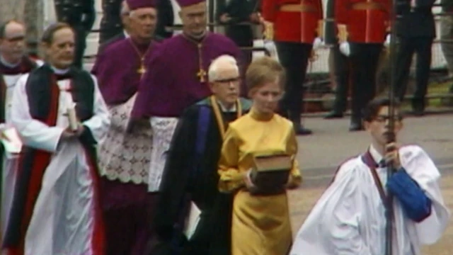 A young Kenner Jones carries a pole with an out of sight cross at the top of it. He is wearing choirboy clothing and glasses. He is leading a procession with a woman, three priests and two bishops.