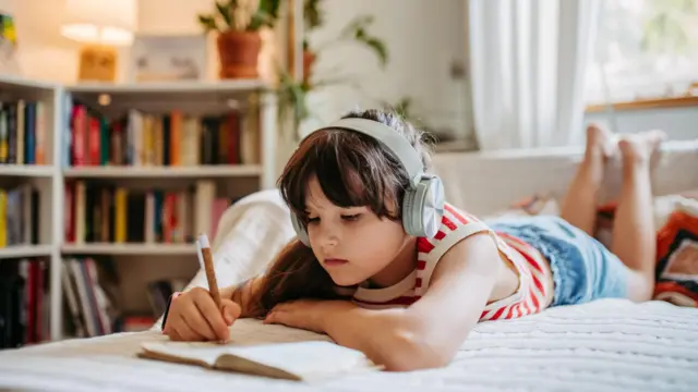 Menina deitada na cama, de fone de ouvido, escrevendo em caderno.