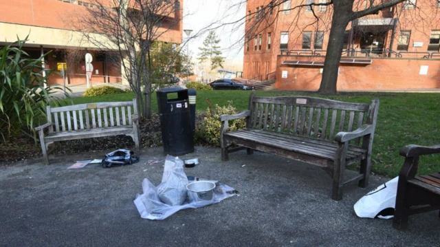 Área no lado de fora do hospital, com três bancos de madeira sobre o cascalho. Uma panela de pressão sem tampa está no chão e sacos estão espalhados ao redor.