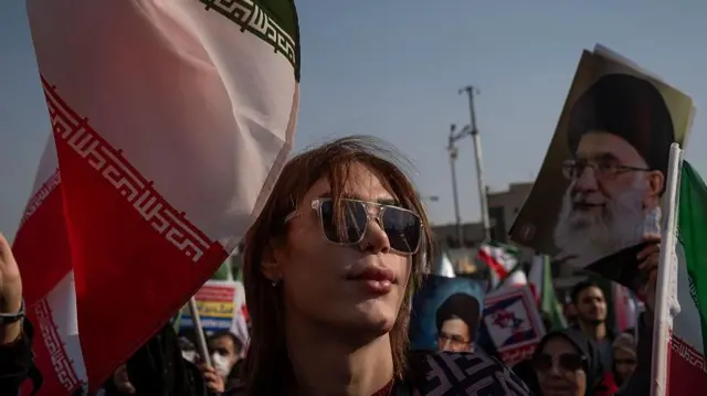 İran bayrağı yanında güneş gözlüklü başı açık bir kadın var. Yanda Hamaney'in bir fotoğrafı duruyor.