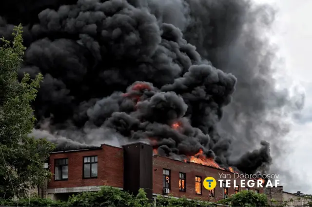Пожежа охопила будівлю, над якою здіймаються густі клуби чорного диму. З даху та вікон виривається полум’я, навколишню територію частково затягує димова завіса.