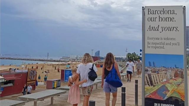 Na orla, três pessoas passam próximas a painel com publicidade que diz em inglês: 'Barcelona: nossa casa. E sua'