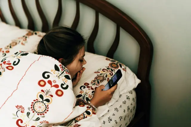 Młoda kobieta leży w łóżku, w pościeli zdobionej kwiatowym wzorem. Ziewa i patrzy w ekran smartfona. 