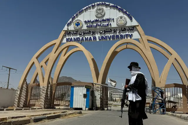 Мужчина с автоматом у ворот, на которых написано Kandahar University.