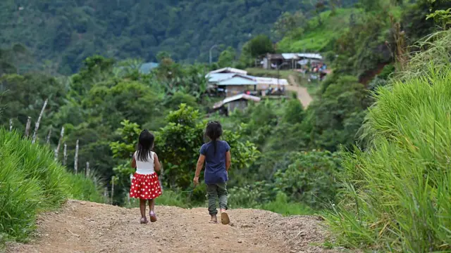 两名年幼儿童均留着长发，正沿着一条泥土小路前行，四周被灌木和绿色植被环绕。其中一人身穿带有白色圆点的红色裙子和粉红色鞋子；另一人身穿蓝色T恤、绿色长裤和人字拖。在远处下方，可见几幢屋顶看似由波纹铁皮搭建的建筑物。