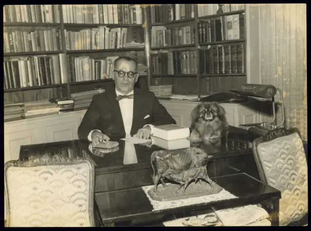 Guimarães Rosa em seu escritório, com um cachorro em cima da mesa e uma estante de livros ao fundo