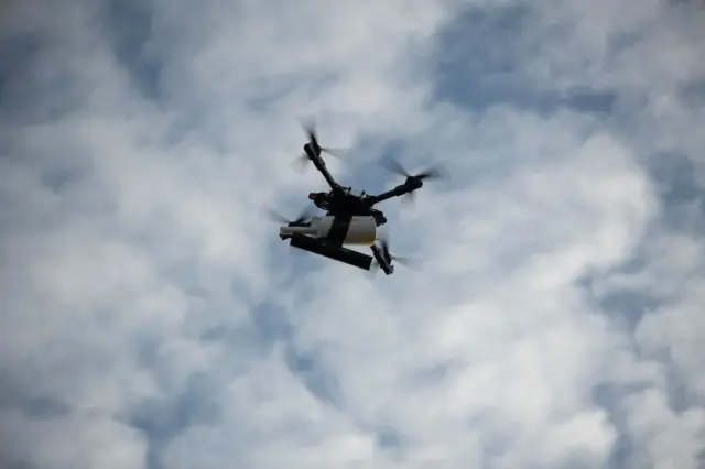 Квадрокоптер, що летить у небі на тлі білих хмар. Він несе під собою скид, а за ним тягнеться напівпрозорий кабель