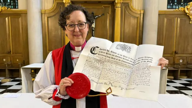 Bishop of St Edmundsbury and Ipswich, Dr Joanne Grenfell, holds a legal document inside a church. She is dressed in her bishop's robes. She has short black hair, glasses and she is smiling at the camera.