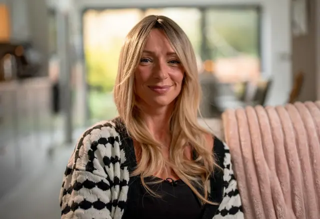 Ellen, uma mulher de longos cabelos loiros, está sentada em um sofá sorrindo para a câmera