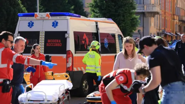 Paramedics in red uniforms attend to injured people, an ambulance can bee seen as well as a stretcher in addition to civilians