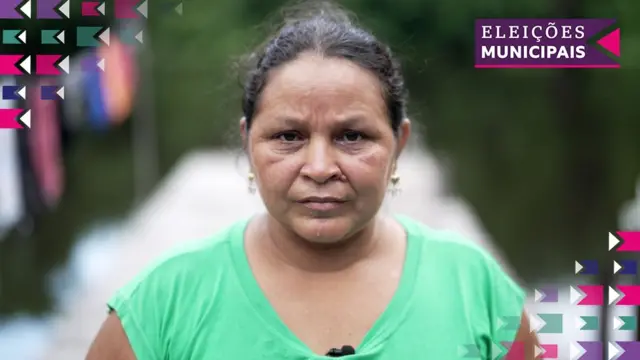 Mulher com camiseta verde, pele morena e cabelos lisos olhando para a câmera; atrás dela há um rio, um varal com roupas e uma floresta ao fundo.