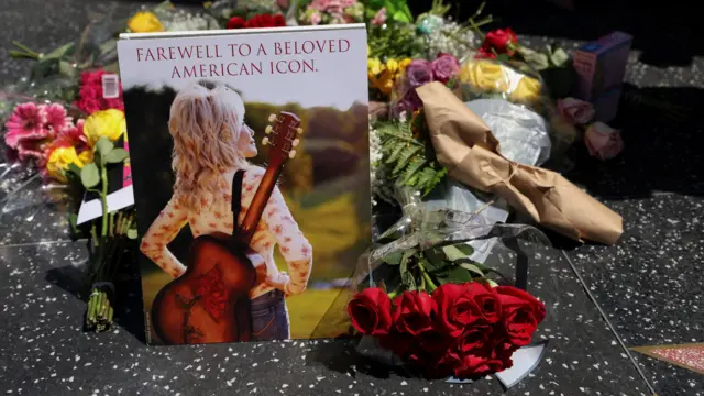 2026年8月25日，在美国加州洛杉矶，乡村音乐传奇人物 Dolly Parton 逝世，终年80岁的消息公布后，人们在她位于荷里活星光大道上的星形纪念章前献上鲜花。花束左侧竖立了一张 Dolly Parton 背着吉他，背对镜头，望向阳光的照片，照片顶部印上字句：“告别广受爱戴的美国传奇。”