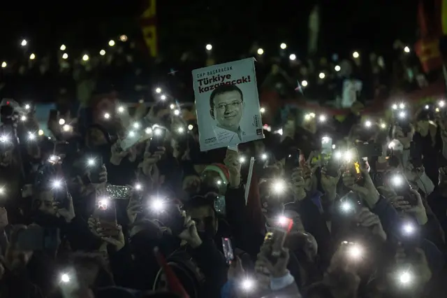 Gece toplanmış bir kalabalık, telefonlarının ışıkları açık, ortada bir kişi üzerinde İmamoğlu'nun fotoğrafı olan ve "Türkiye kazanacak" yazan bir pankart tutuyor. 