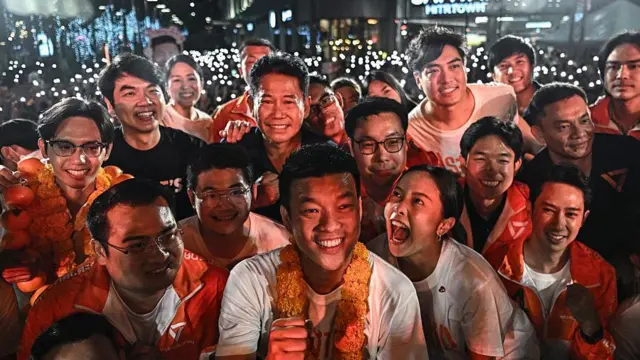 Young Thai voters wearing white shirts and orange jackets, holding up their phones as lights at an election campaign rally