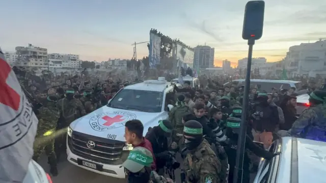 A Red Cross vehicle is surrounded by a crowd of people including photographers and Hamas fighters