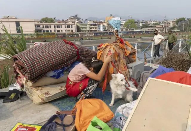सुकुम्बासी बस्तीमा घरायसी समानहरू बीच कुकुर समातेर बसेकी एक बालिका