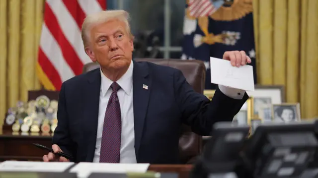 Donald Trump hold up a letter as e sit for im desk for di Oval Office holding a pen.