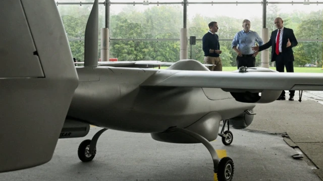 A military drone stands in the foreground - about the height of a person and in the shape of a plane. Three men, including Defence Secretary John Healey stand in the background of the hangar, which has large plate-glass windows at the back, looking onto greenery.