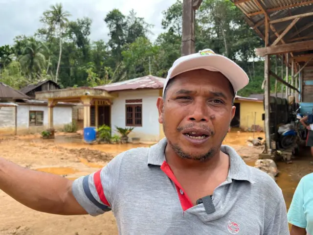 Anwar Pangabean menggunakan topi, dan kaos berkerah di latar belakang rumah terdampak banjir. 