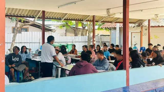 Hàng chục sinh viên đang ngồi trong lớp học, trong khi một người đàn ông đứng ở phía trước