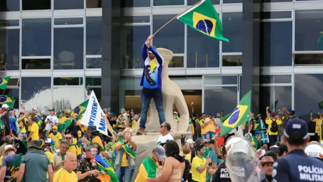 Centenasmines onabetpessoasmines onabetvoltamines onabetestátua da Justiçamines onabetfrente ao STF; há um manifestantemines onabetpé,mines onabetcima da estátua, erguendo bandeira do Brasil