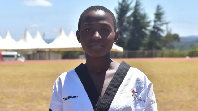 Laurence Namukiza, vêtu d'une tenue de taekwondo blanche avec des bordures noires, regarde la caméra alors qu'une grande tente et des arbres sont visibles à l'arrière-plan.