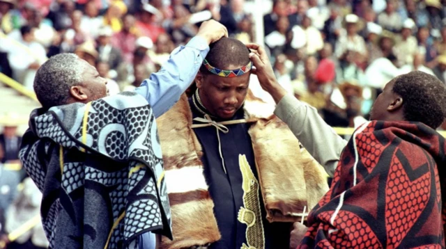 Two traditional chiefs put a calfskin headband and feather on the new monarch's head