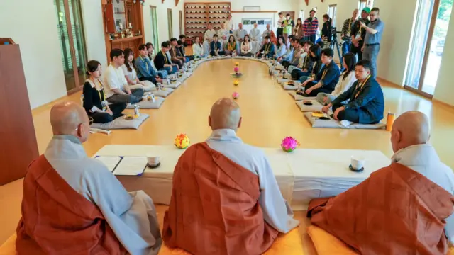 Un numeroso grupo de participantes está sentado sobre cojines colocados en un amplio semicírculo en el interior de la sala de un templo, con tres monjes al frente.